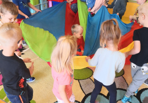 dzieci na dywanie stoją z kolorową chustą animacyjną trzymają jej brzegi, na niej siedzi jedno z dzieci